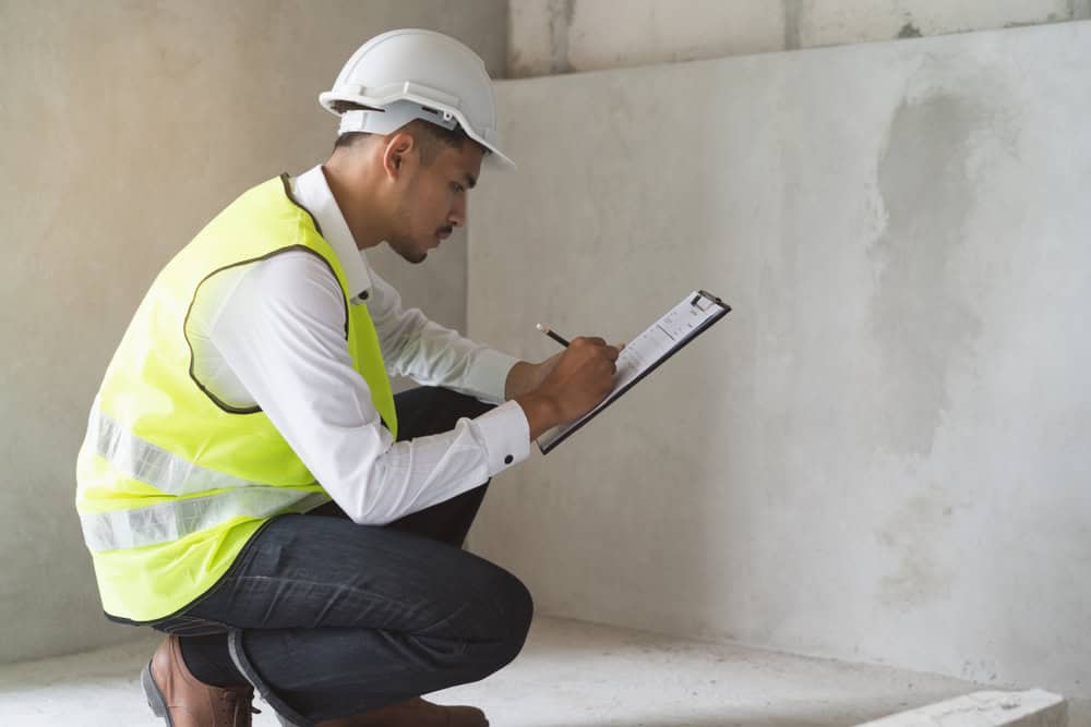 A construction worker in a safety vest and helmet takes detailed notes on a clipboard at a building site.
