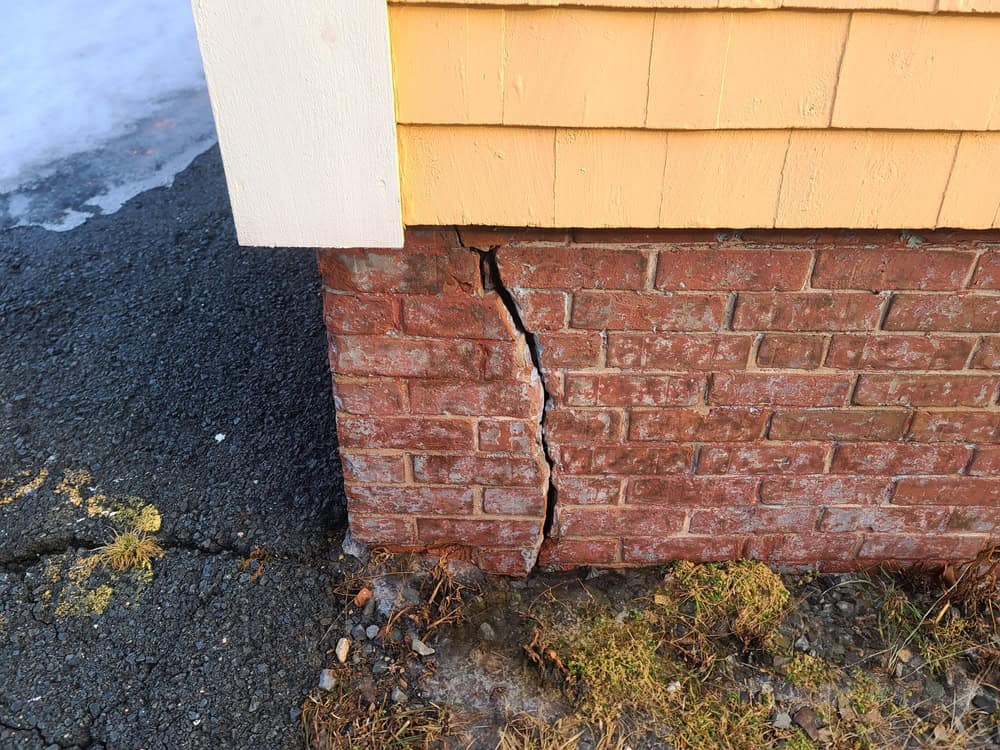 A close-up of a cracked brick foundation under a yellow house, hinting at potential structural issues.
