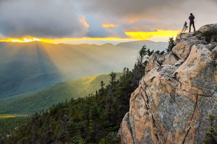A lone hiker stands atop a dramatic cliff edge, silhouetted against a spectacular mountain sunset as golden rays pierce through stormy clouds in the Adirondack wilderness.
