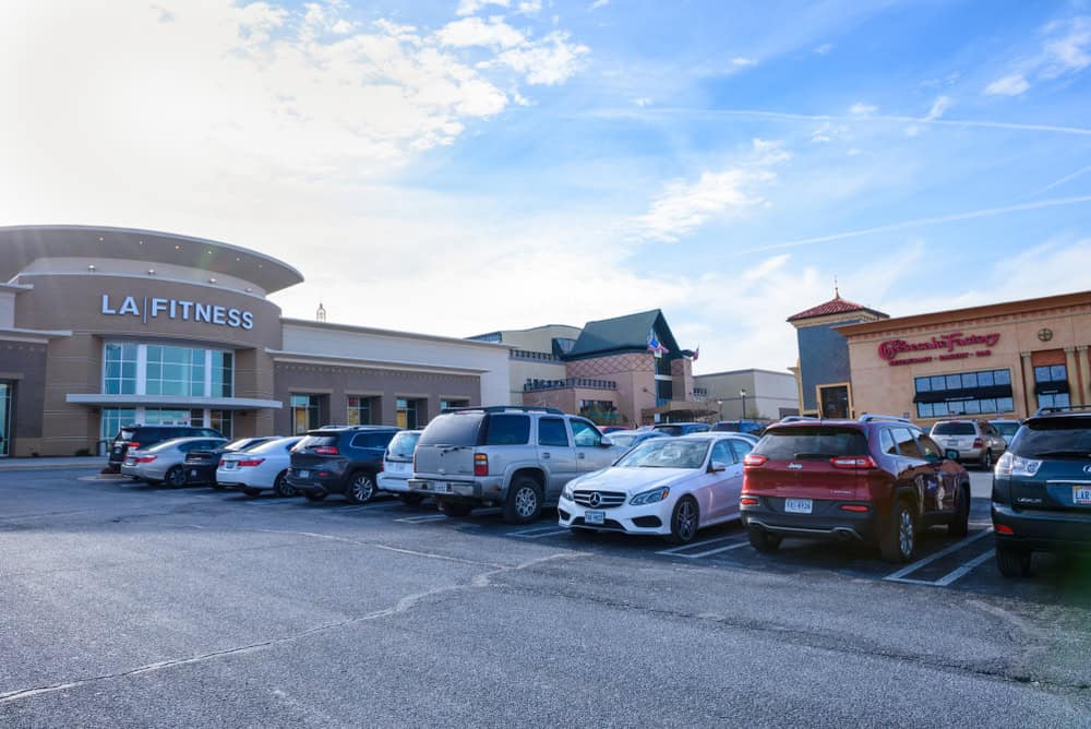 A bustling strip mall parking lot on a bright day, anchored by LA Fitness, with a row of cars parked outside various retail storefronts under wispy blue skies.