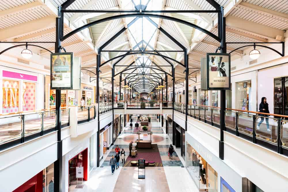 Modern mall elegance: Arched ceilings and black beams frame a bustling two-story shopping center, where vibrant storefronts and digital displays line the walkways as shoppers explore the spacious, light-filled interior.