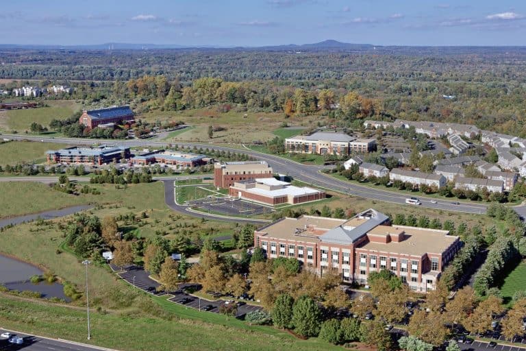 Aerial view of a thriving suburban community showcasing modern buildings, residential areas, and expansive green spaces, with forested hills stretching to the horizon.