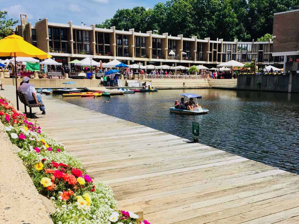 Lakeside relaxation: A vibrant summer scene at Lake Anne Plaza in Reston, Virginia, complete with colorful market umbrellas, paddle boats, and blooming flower beds along the wooden boardwalk.