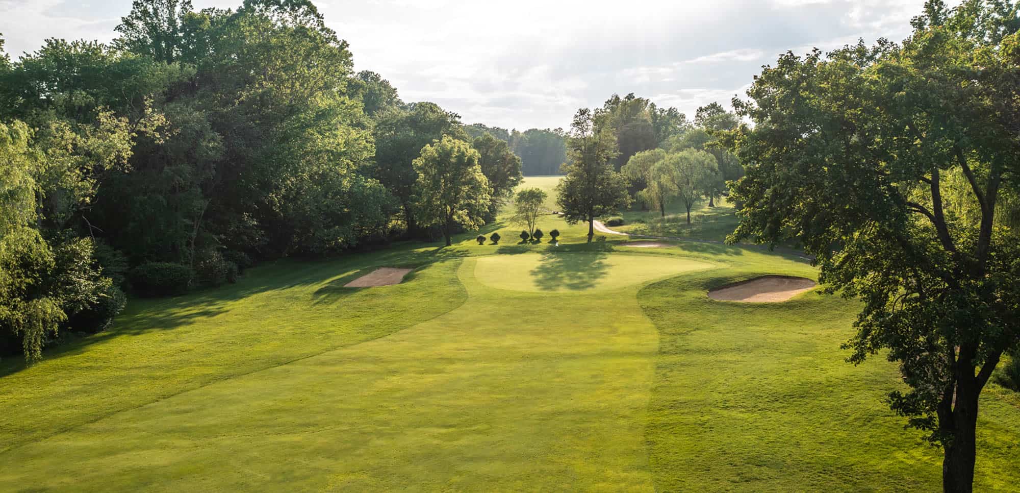 A pristine golf hole winds through mature trees, featuring manicured fairways, strategic bunkers, and an elevated green bathed in warm evening light.