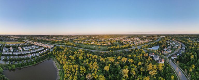 A serene aerial view of a sprawling suburban neighborhood bordered by lush greenery and ponds under a clear blue sky.