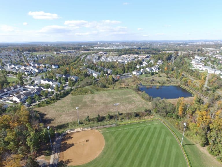 Suburban sprawl meets nature: Aerial view of a residential neighborhood with baseball diamond, open fields, and a serene pond, showcasing the blend of community amenities and green spaces.