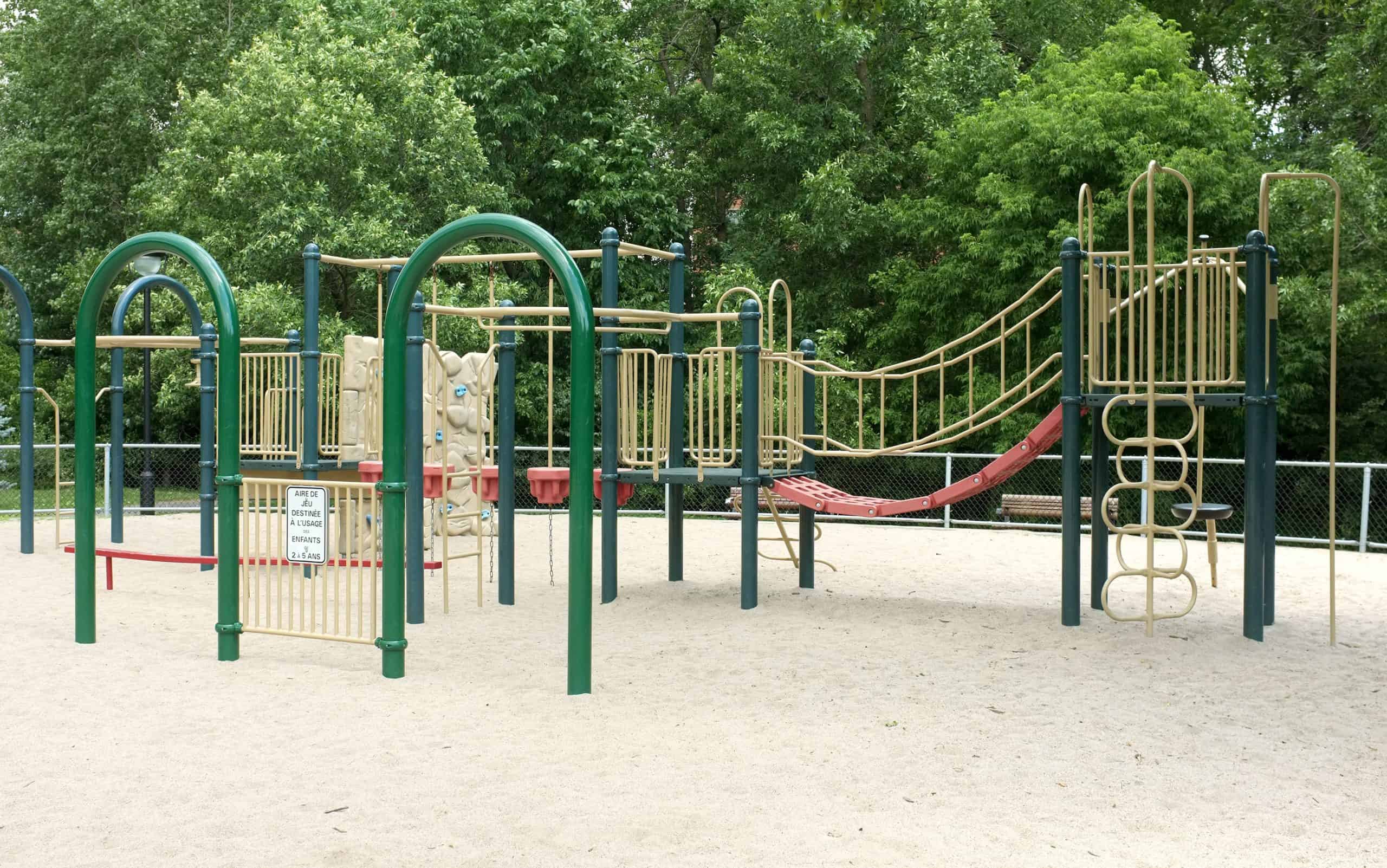 Modern playground equipment featuring green arches, climbing structures, and a red slide set against a backdrop of lush summer foliage