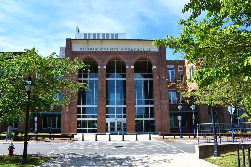 Fairfax County Courthouse stands proudly with its distinctive brick facade and soaring glass windows, framed by leafy green trees on a bright summer day.