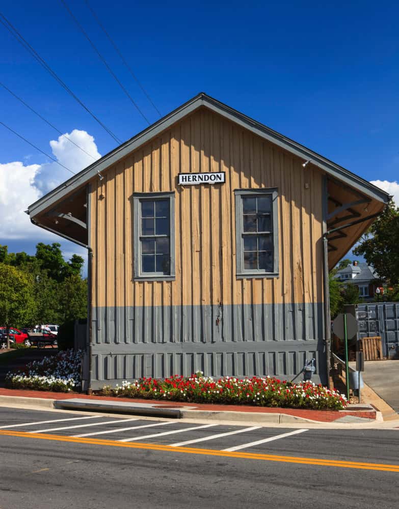 Historic Herndon train depot with its distinctive yellow and gray board-and-batten siding, adorned with vibrant flower beds beneath a bright Virginia sky.