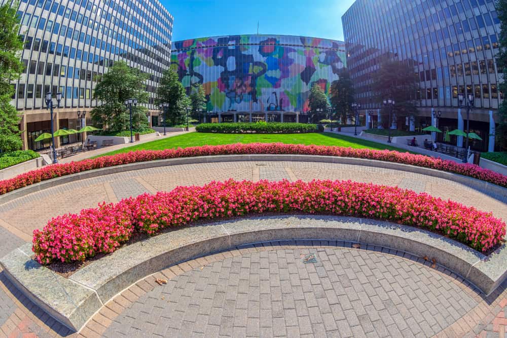 Urban Oasis: Vibrant flower beds frame a modern plaza, with colorful street art adorning a cylindrical building between sleek office towers.