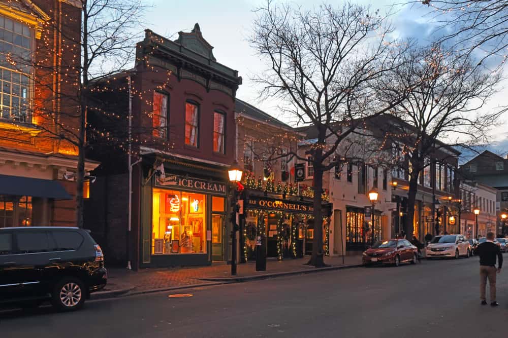 Alexandria VACharming winter evening on a historic street: Twinkling lights adorn storefronts, including an inviting ice cream parlor and O'Connell's pub, as dusk settles over the quaint brick buildings and bare-branched trees.