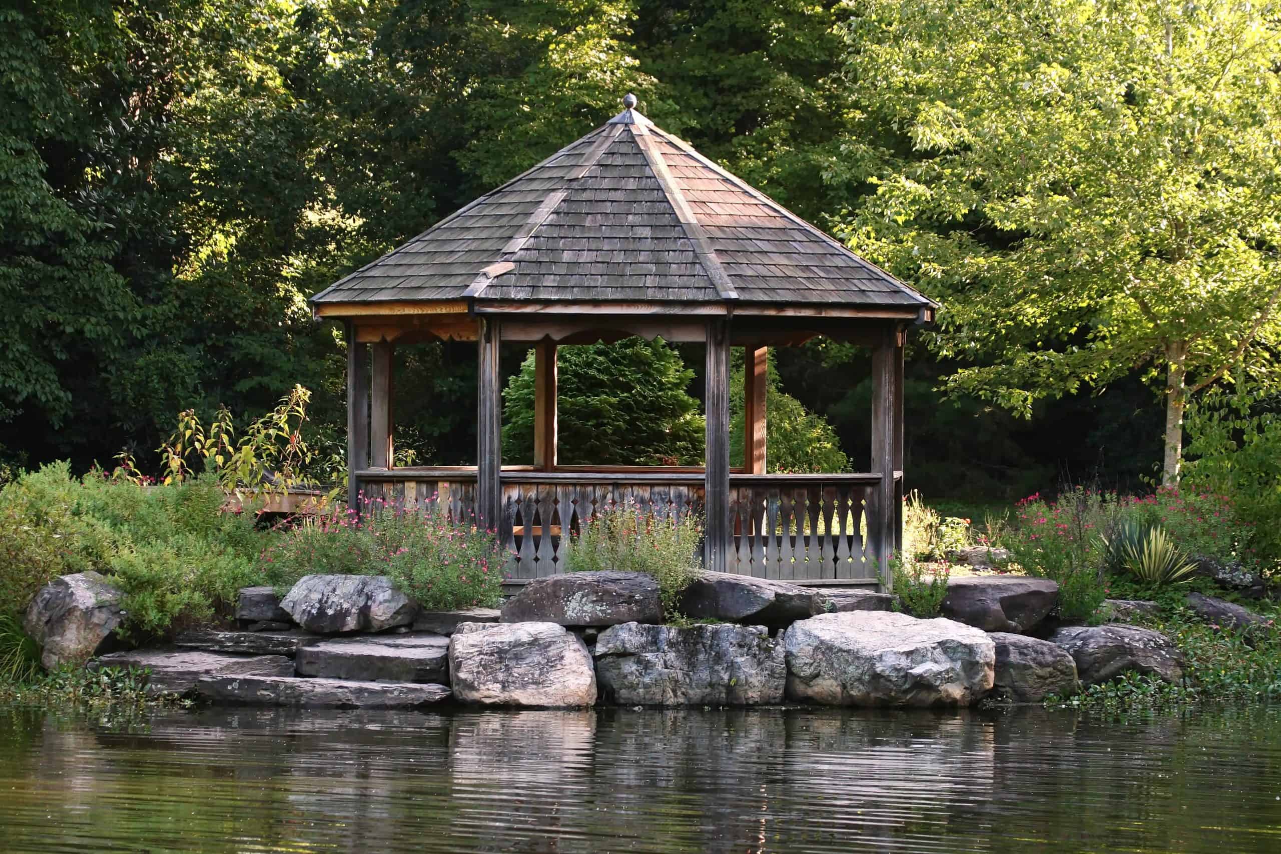 Tranquil waterside gazebo surrounded by wildflowers and rocky shoreline, nestled in a leafy garden setting
