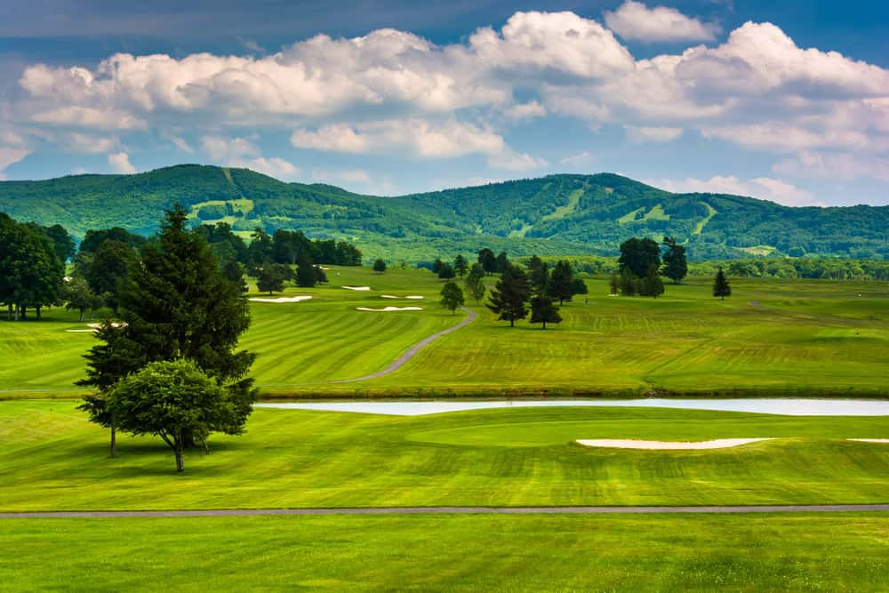 Pristine golf course nestled in the Appalachian Mountains, featuring manicured fairways, white sand bunkers, and ski slopes visible on the verdant summer mountainsides.