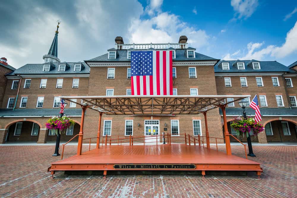 Alexandria VAPatriotic display at the Wiley F. Mitchell Jr. Pavilion: A grand brick building adorned with a massive American flag, flanked by a wooden stage and flowering planters under a bright blue sky.