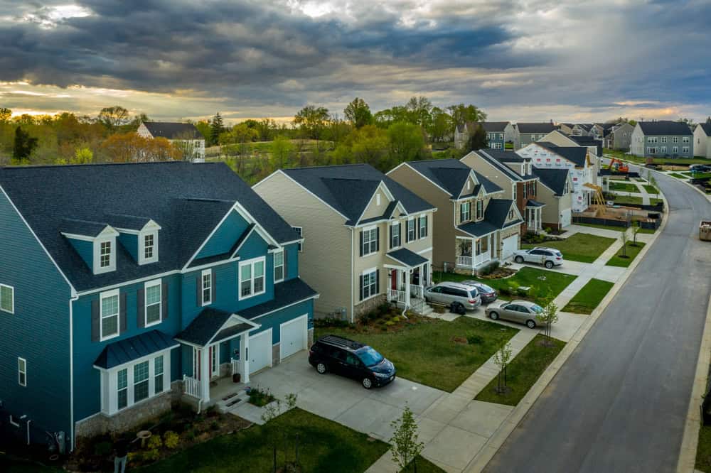 A suburban neighborhood at dusk, with rows of colorful houses along tree-lined streets and cars parked in the driveways under a dramatically cloudy sky.