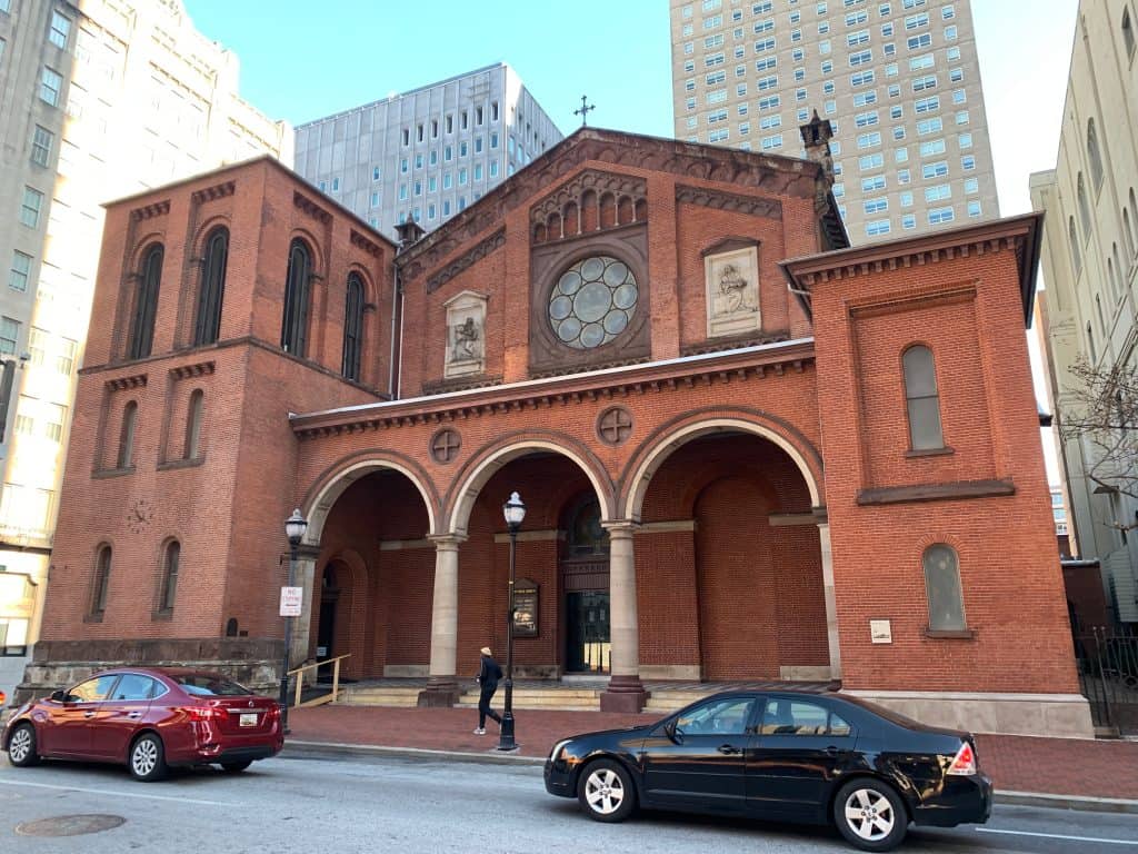 Stately brick church standing tall amidst the urban landscape, with passing cars and a pedestrian highlighting its presence.