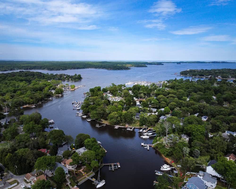 Coastal Living Paradise: Aerial view of a picturesque waterfront community nestled among lush trees, featuring winding waterways, private docks, and a bustling marina, all under a clear blue sky.