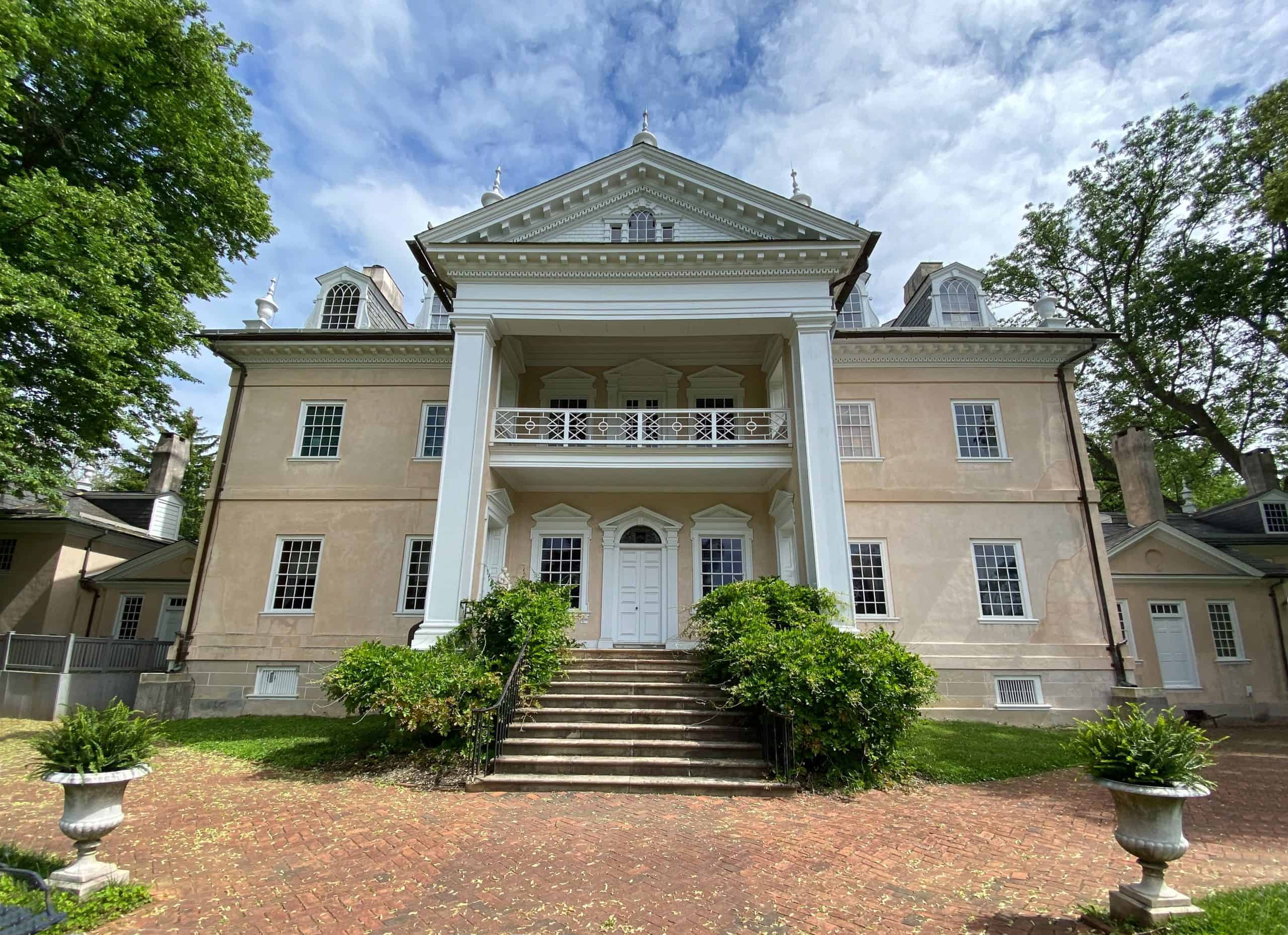 A grand neoclassical mansion stands under a blue sky, framed by lush greenery and stately columns.
