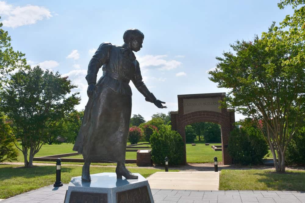 Statue of a woman in front of the Carnegie Building archway on a sunny day, surrounded by lush greenery.