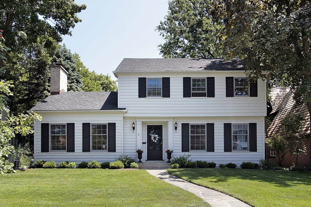 Charming two-story white house with black shutters, nestled amidst lush greenery.