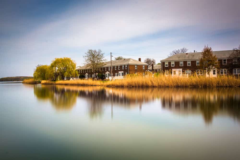 Tranquil riverside rowhouses reflected in serene water, surrounded by lush greenery.