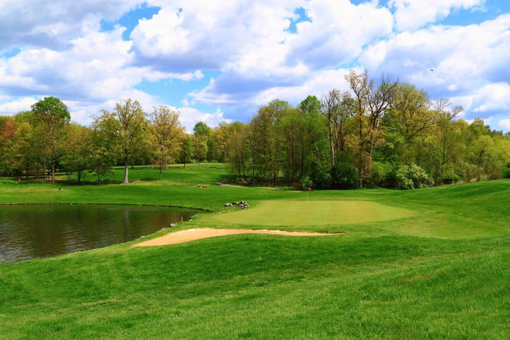 A picturesque golf course on a spring day, featuring a lush green fairway, a small pond, and a sand trap, all framed by budding trees and a partly cloudy blue sky.