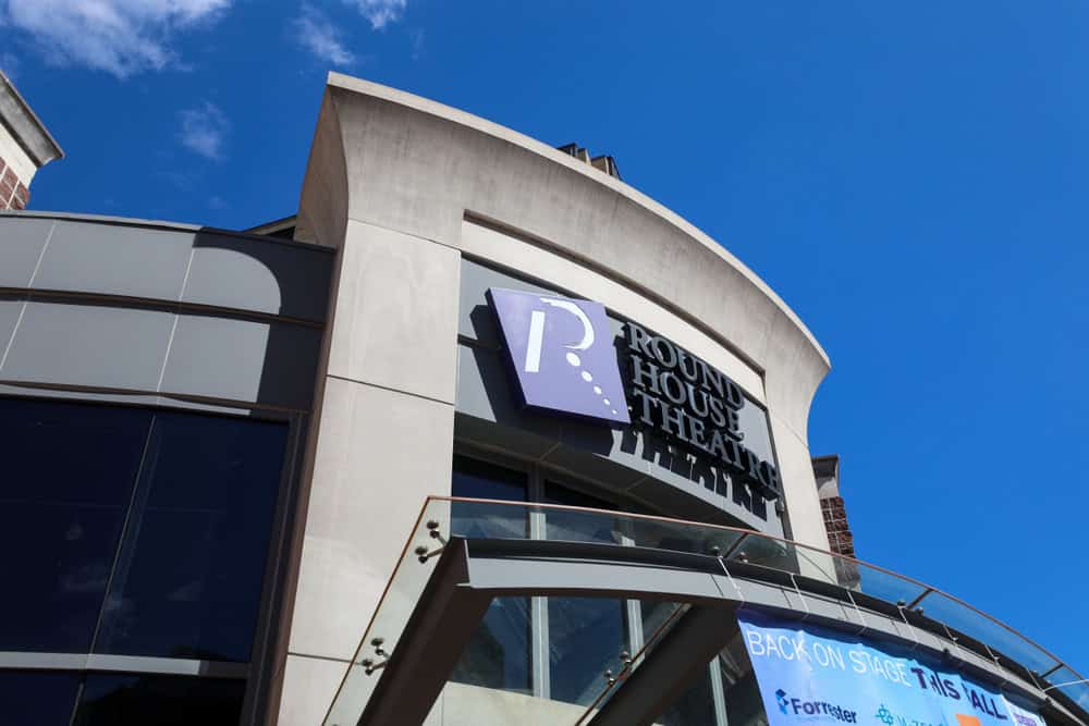 Curved modern architecture of the Round House Theatre entrance against a bright blue sky, featuring the theatre's distinctive purple logo and signage.