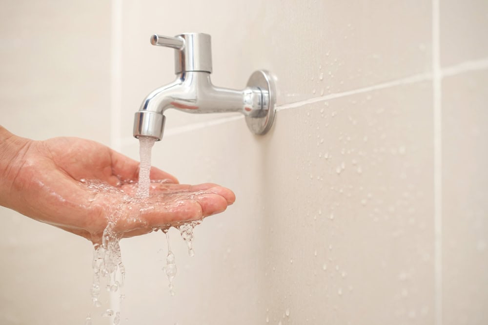 Fresh water flowing from a chrome wall-mounted faucet into a cupped hand, with water droplets splashing against light beige tiled bathroom wall.