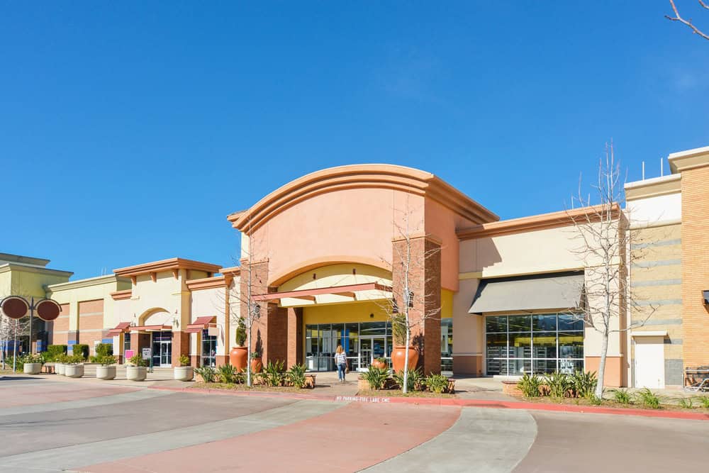 A vibrant shopping center under a clear blue sky, with a welcoming entrance and lush greenery.