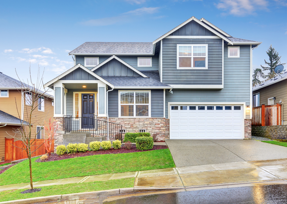 Modern blue suburban home with a charming front porch, two-car garage, and well-manicured yard under a clear sky.