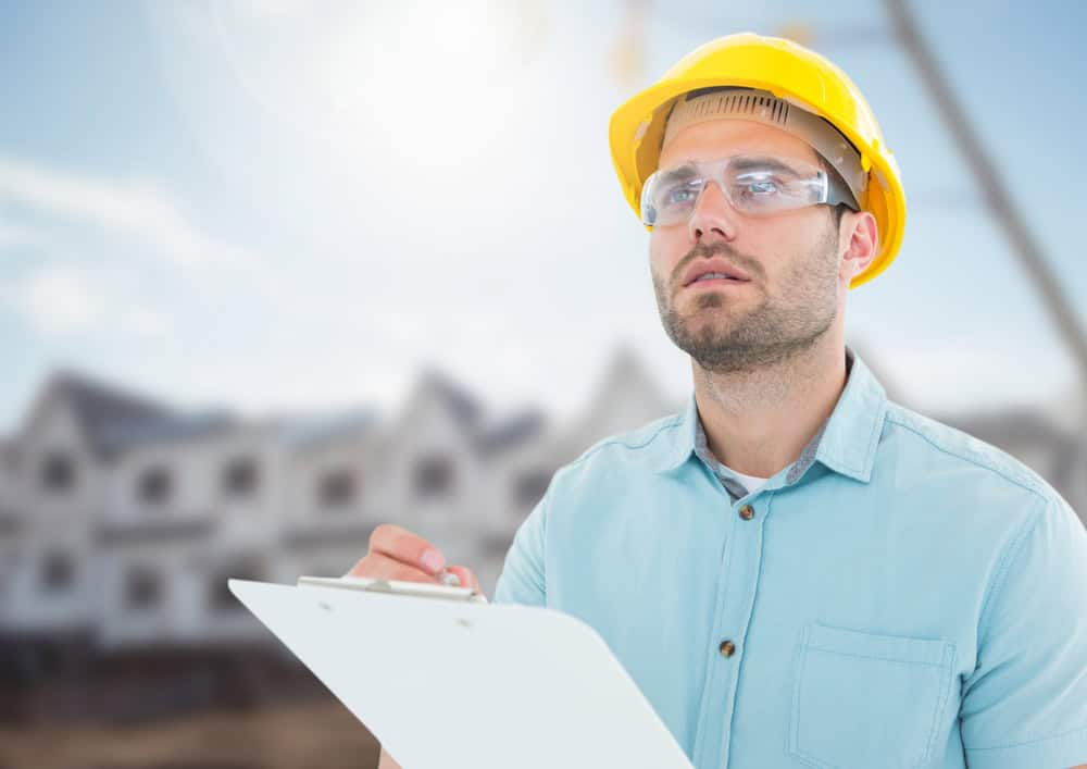 Construction site supervisor in a yellow hard hat inspects progress, clipboard in hand, against the backdrop of new houses under a bright sky.