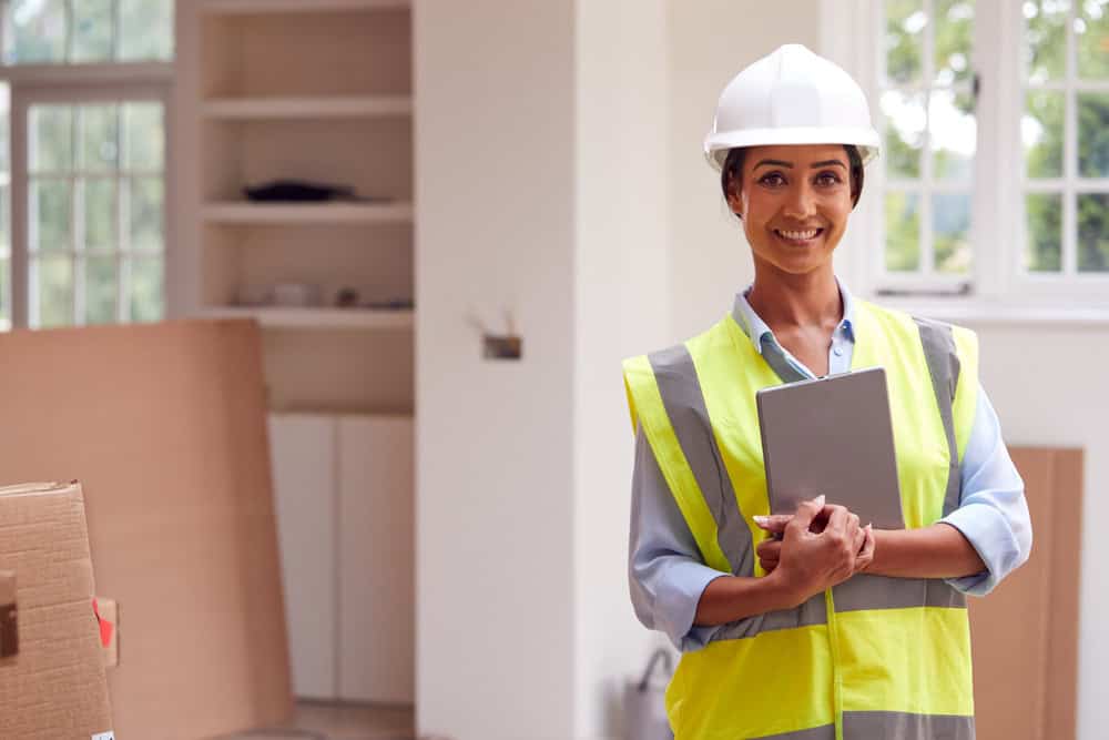 Confident female construction manager in a hard hat and safety vest stands with a clipboard inside a renovation project.