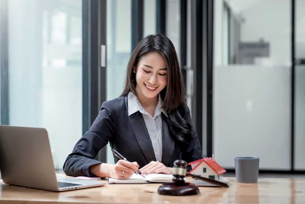 A smiling businesswoman at her desk, surrounded by a laptop, gavel, and miniature house, working in a modern office.
