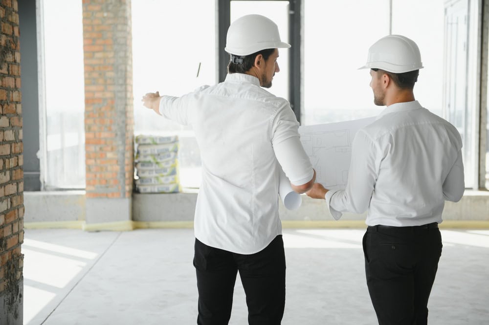 Two architects in hard hats discuss building plans inside a construction site.