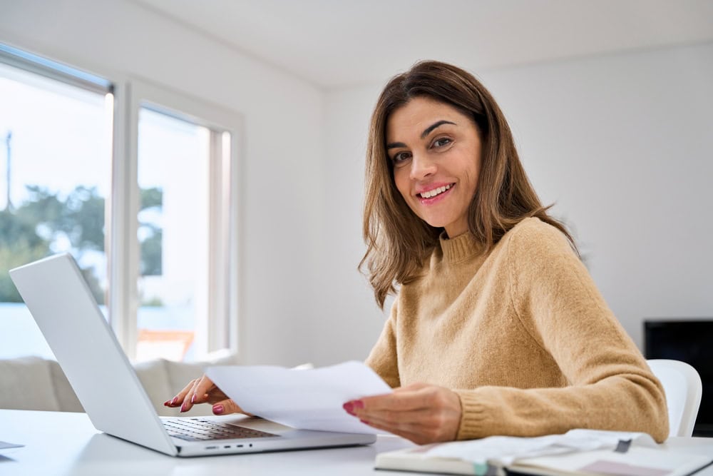 Smiling woman working from home at her laptop, reviewing documents in a bright, modern room.