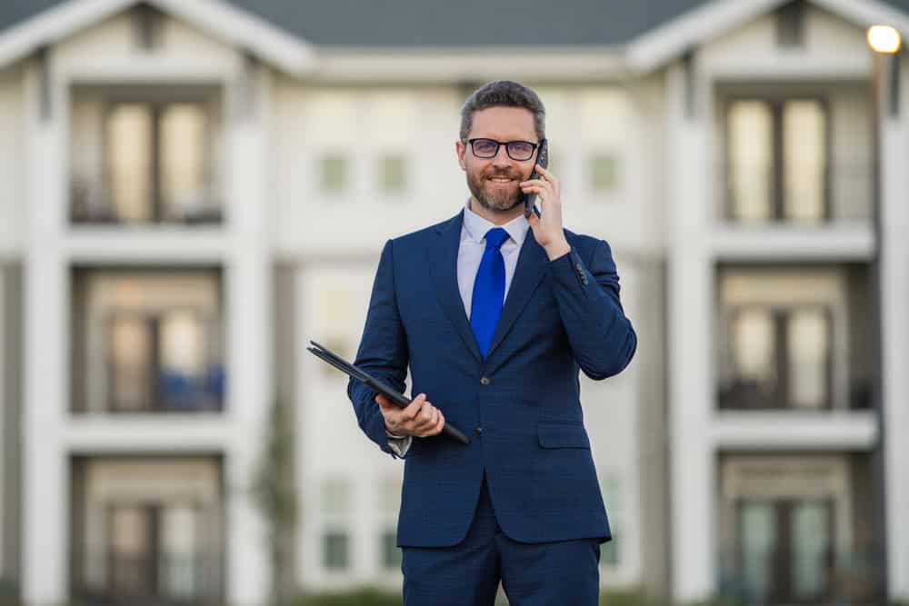Confident real estate agent in a blue suit stands outside modern apartment complex, talking on the phone and holding a tablet.
