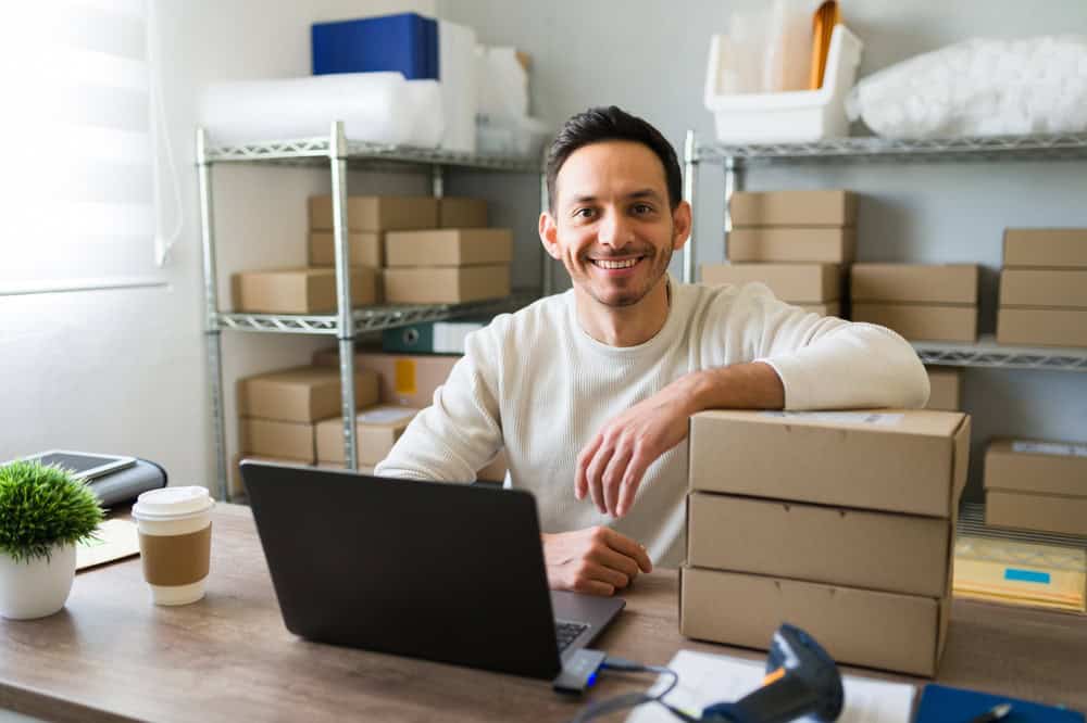 Entrepreneur smiling at his desk in a small business warehouse, surrounded by shipping boxes and ready to fulfill online orders.