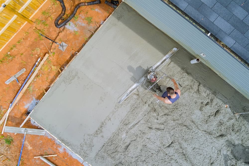 Aerial view of a construction worker using a power screed to level a fresh concrete slab beside a house.