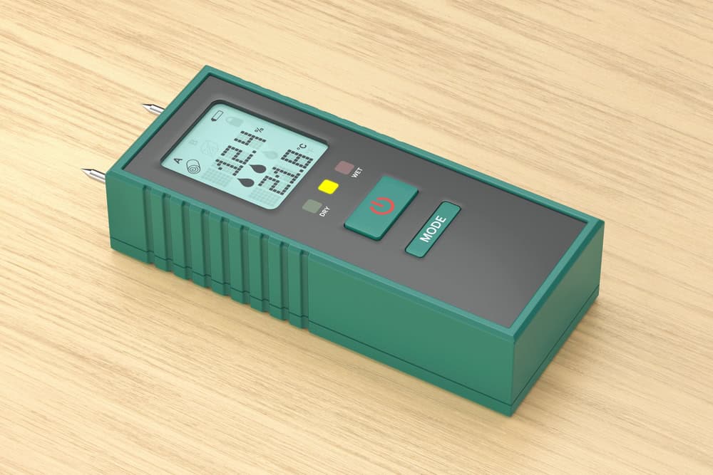 Green handheld moisture meter with LCD readout and dual probe pins resting on a wooden surface.