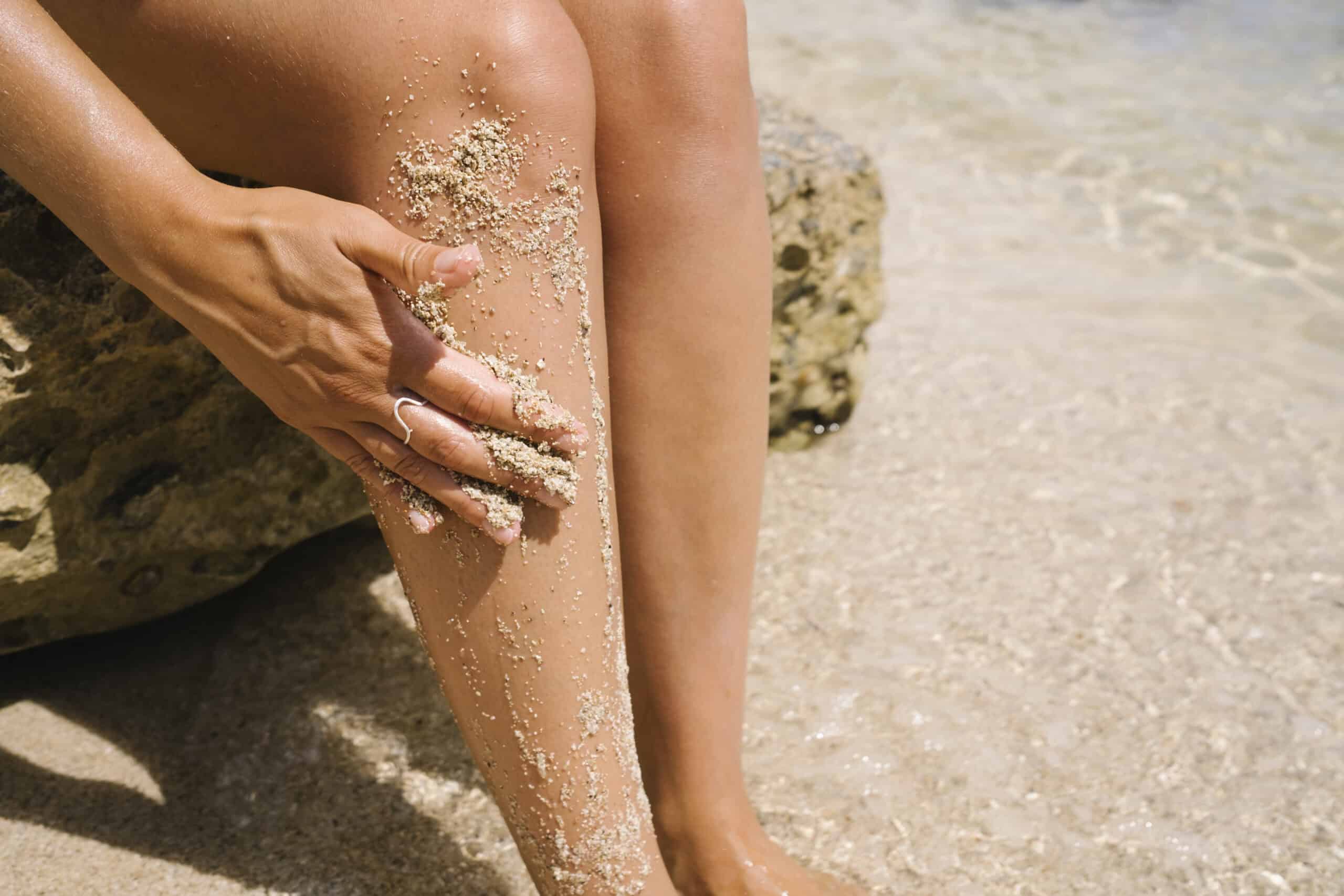 Woman exfoliating legs with sand sitting on rock at beach