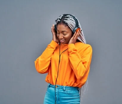 Adorable afro american woman in yellow shirt singing and having fun while listening to music using earphones isolated over gray background. Copy space