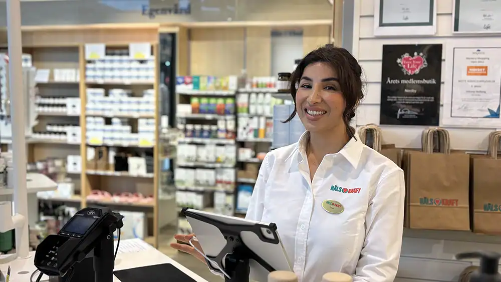Kvinne i helsebutikk smiler foran kasse og produkter.