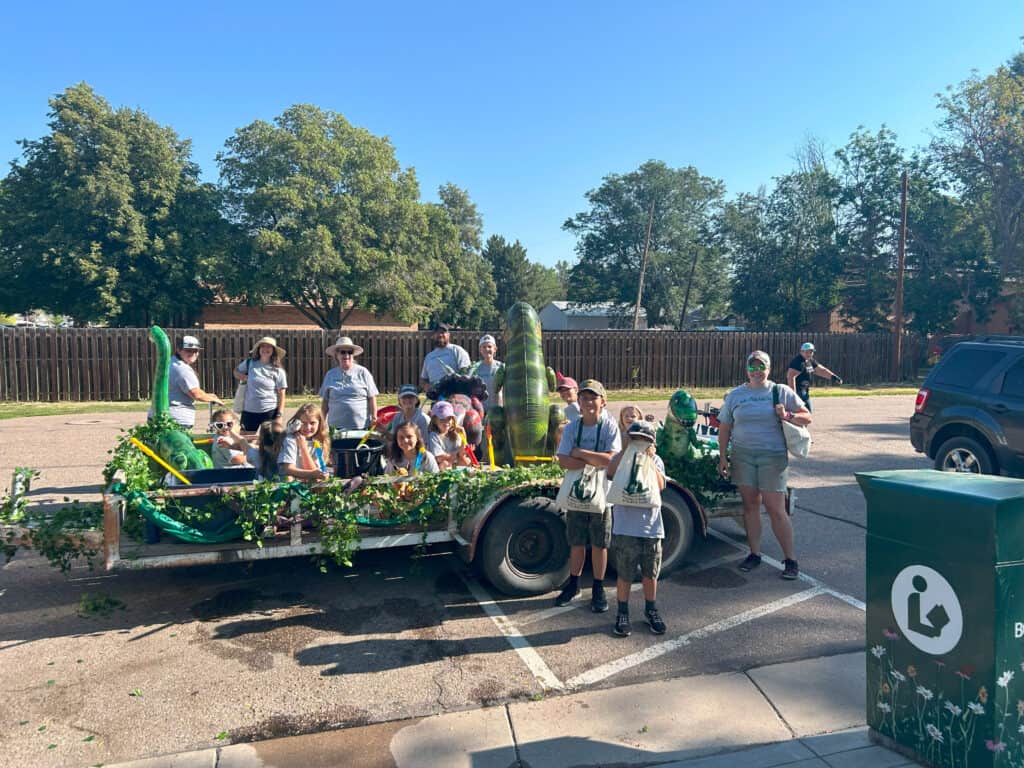 Colorful float decorated with green plants and dinosaur inflatable figures, featuring children and adults participating in a parade or community event on a sunny day in a neighborhood.