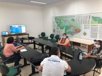 Conferência sobre políticas ambientais e gestão territorial com representantes ambientais em sala de reunião, usando máscara, com mapas e telas digitais relacionadas à sustentabilidade e meio ambiente.