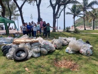 Enterrro de lixo acumulado em área verde de praia com equipe de voluntários ao fundo.