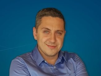 Homem sorridente com camisa azul escura em fundo azul, representando profissionalismo e confiabilidade na área de notícias ambientais e mercantis.