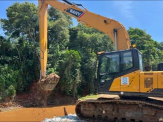 Máquina de escavadeira em operação na construção civil, removendo terra de área de floresta com vegetação densa ao fundo.