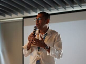 Imagem de um homem falando em um evento de sustentabilidade e meio ambiente, com microfone na mão, usando óculos e camisa social, diante de tela de projeção branca, promovendo conscientização ambiental e estratégias sustentáveis.