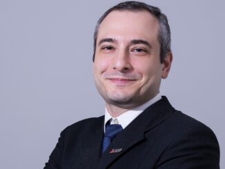 Imagem de um homem de terno preto, camisa branca e gravata azul, sorrindo confiantemente. Perfil profissional adequado para temas relacionados a negócios, sustentabilidade e meio ambiente. Foco em imagem corporativa moderna.