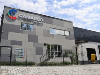 Camargo Química fachada de fábrica moderna com logo visível sob céu nublado, área de entrada com cerca, sinalizações de recepção, expedição e visitantes, representando um ambiente industrial limpo e seguro.